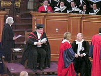 Degree ceremony at the University of Oxford. The Pro-Vice-Chancellor in MA gown and hood, Proctor in official dress and new Doctors of Philosophy in scarlet full dress. Behind them, a bedel, another Doctor and Bachelors of Arts and Medicine.