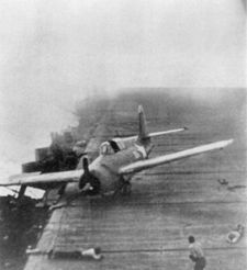 A Hornet Wildcat that just landed minutes earlier skids across Enterprise’s flight deck as the carrier maneuvers violently during Junyo’s dive bomber attack.  Two crewmen are taking defensive postures on the deck as smoke from earlier bomb hits swirls around them.