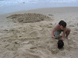 One person is buried in sand by another; their sandcastle is visible in the background.