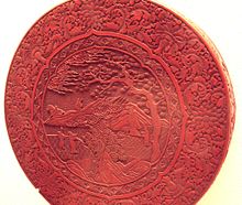 A Ming Dynasty red lacquer box with intricate carving of people in the countryside, surrounded by a floral border design.