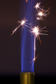 The sparks generated by striking steel against a flint provide the activation energy to initiate combustion in this Bunsen burner. The blue flame will sustain itself after the sparks are extinguished because the continued combustion of the flame is now energetically favorable.